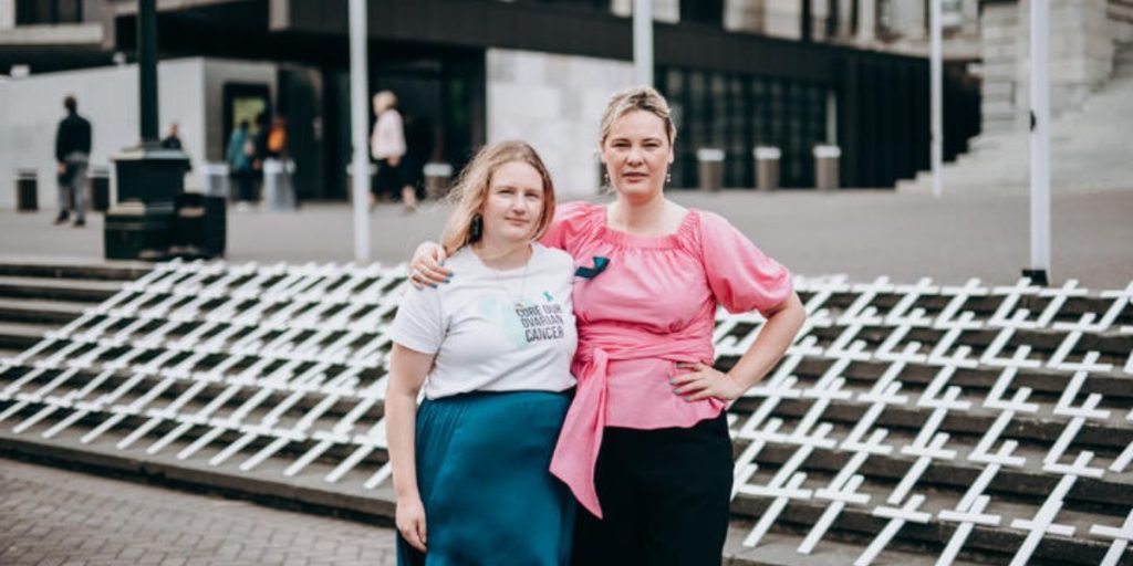 two wmen standing outside parliament in front of white crosses that they have laid on parliament steps to signify the women that have died of ovarian cancer